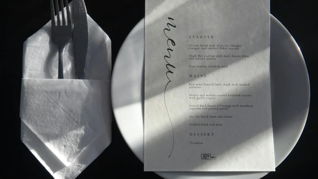 Overhead fine-dining place setting: white plate, folded napkin, and a handwritten menu card.