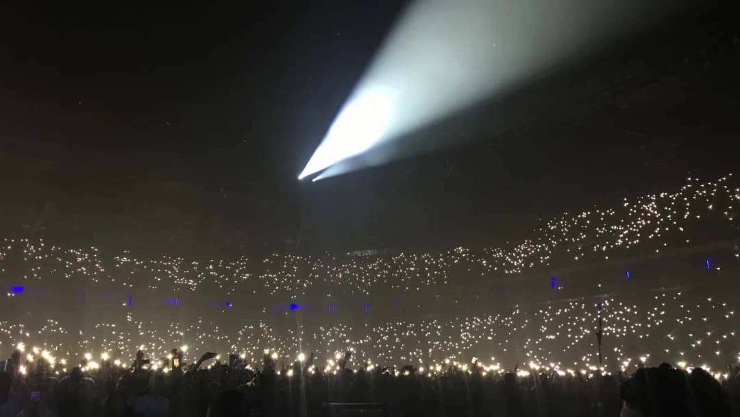 Packed arena crowd glowing with phone lights under a white spotlight at Paris La Défense Arena