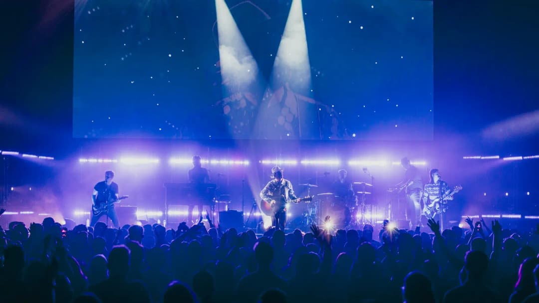 Band on a blue-lit stage in front of a silhouetted concert crowd