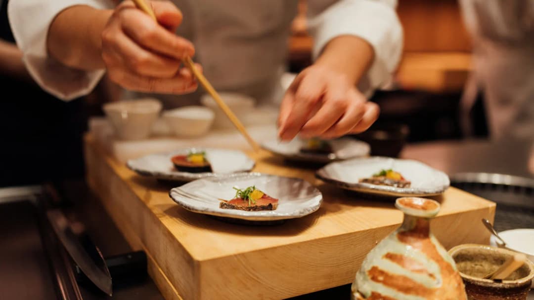 Chef's hands plating small tasting-menu dishes on a wooden board