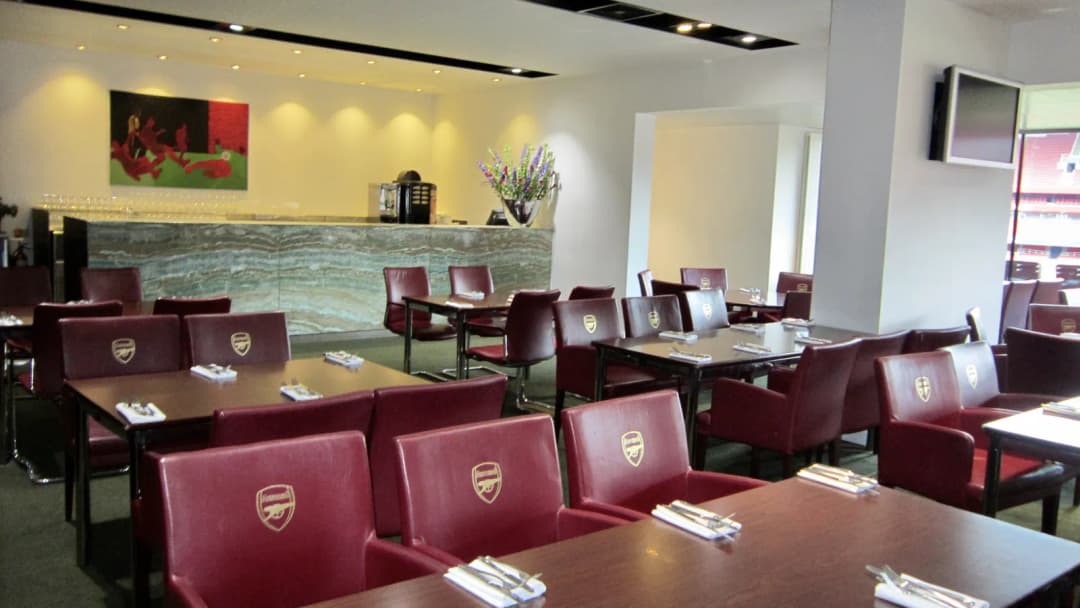Stadium hospitality dining room with red leather chairs bearing club crests, tables laid for guests