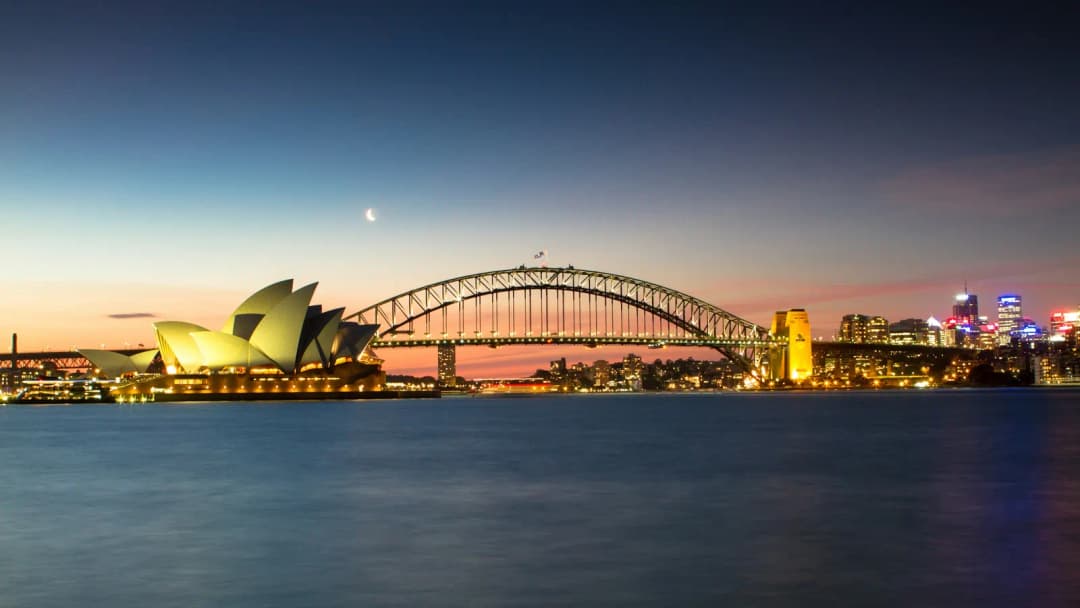 Sydney Harbour at dusk with the Opera House and Harbour Bridge lit against the sky.