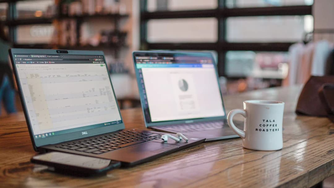 Two open laptops side by side on a wooden desk beside a white coffee mug