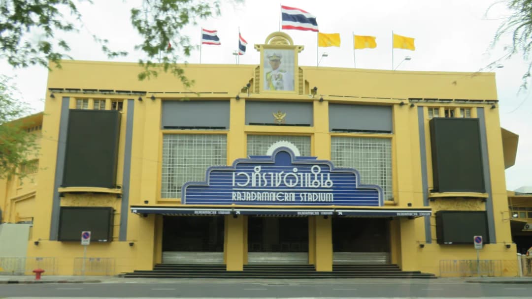 Yellow facade of Rajadamnern Stadium in Bangkok, its name on the entrance sign, flags on the roof.