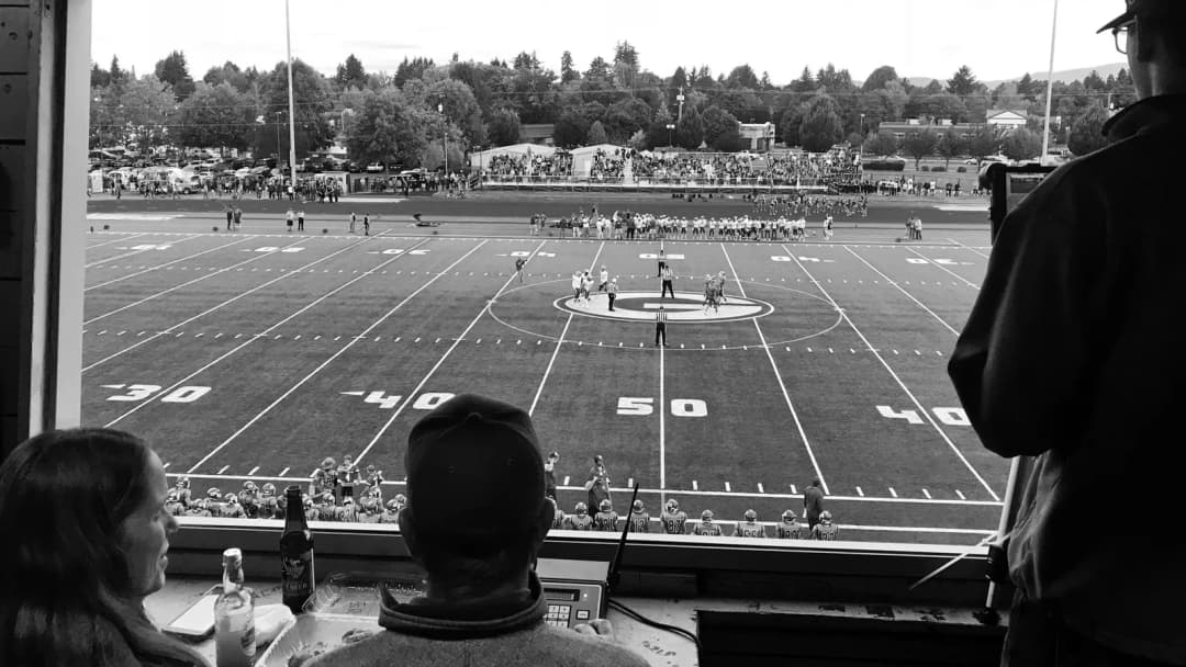Two people watch an American football game from a window overlooking the field, in black and white