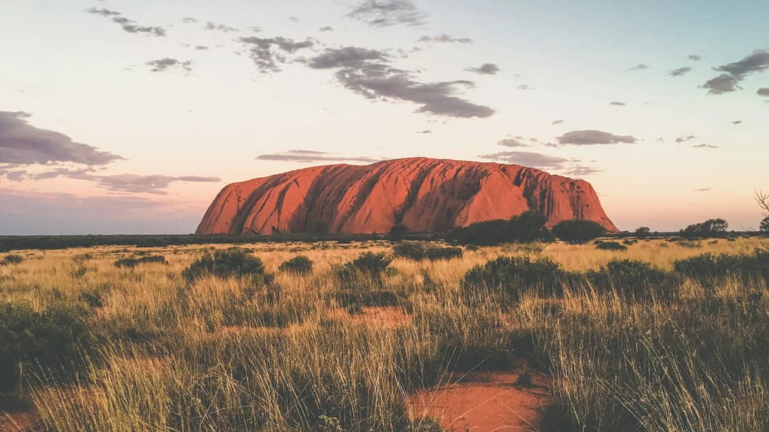 Uluru rising above dry outback grass at sunset, under a pale sky with clouds.