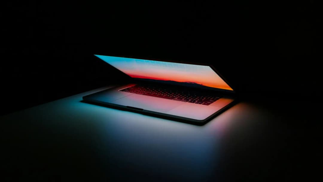 A laptop glowing on a dark desk in a dim room.